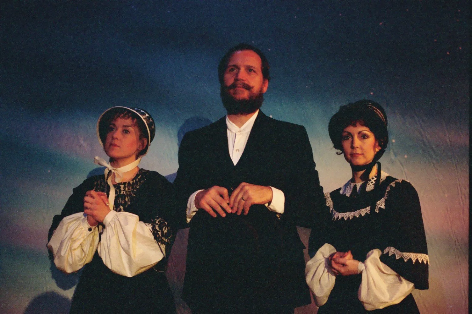 Three people in period costumes, two women in bonnets and a man with a beard, stand against a dark backdrop. One woman is on the left and another on the right, with the man in the center. Both women wear lace-trimmed garments, and the man wears a for