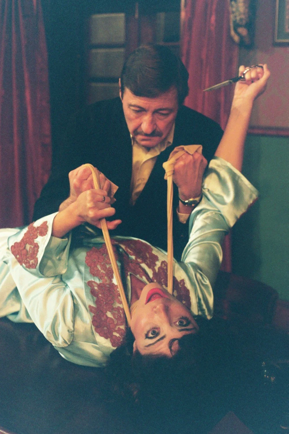 A man holding a woman with long ropes in a dimly lit room. The woman is lying down, wearing a light blue robe with red embroidery. The man is looking down, holding the ropes with scissors in his hand.