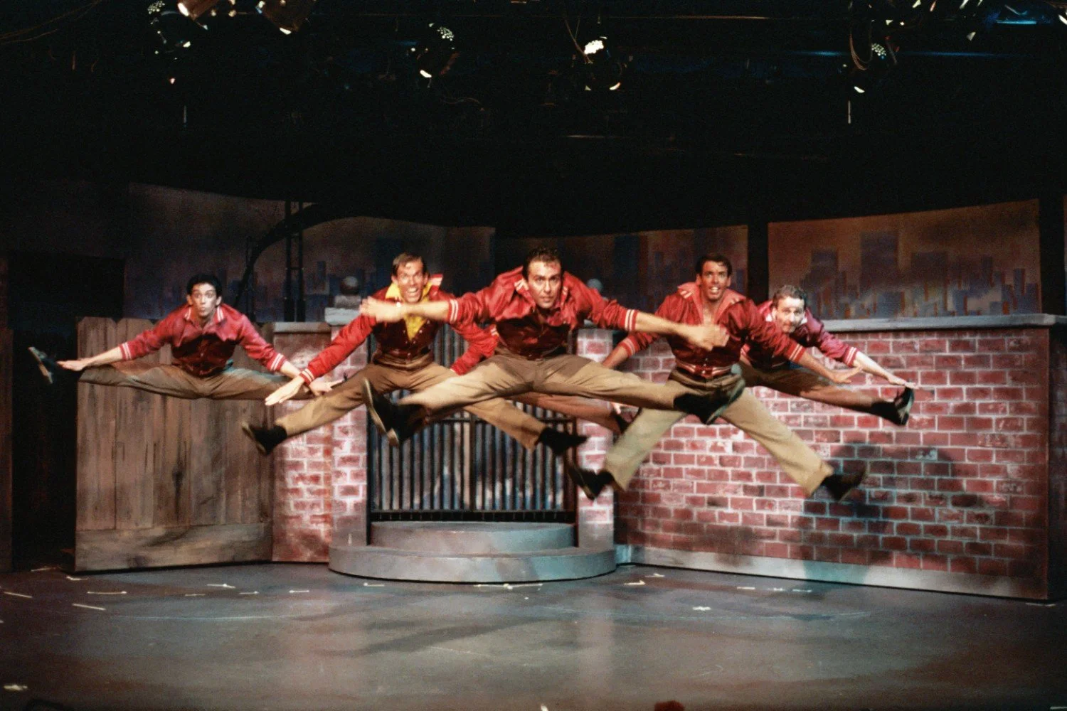 Stage performance with five men in red jackets performing a synchronized leap against an urban backdrop.