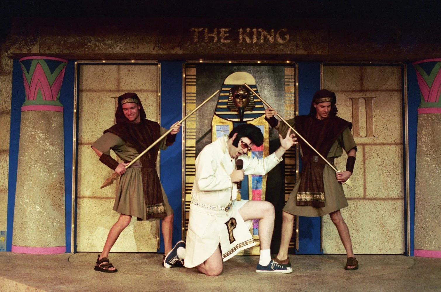 Costumed performer in white with microphone kneeling on stage between two individuals dressed as guards holding spears, in front of an ancient Egyptian themed backdrop with "The King" sign.