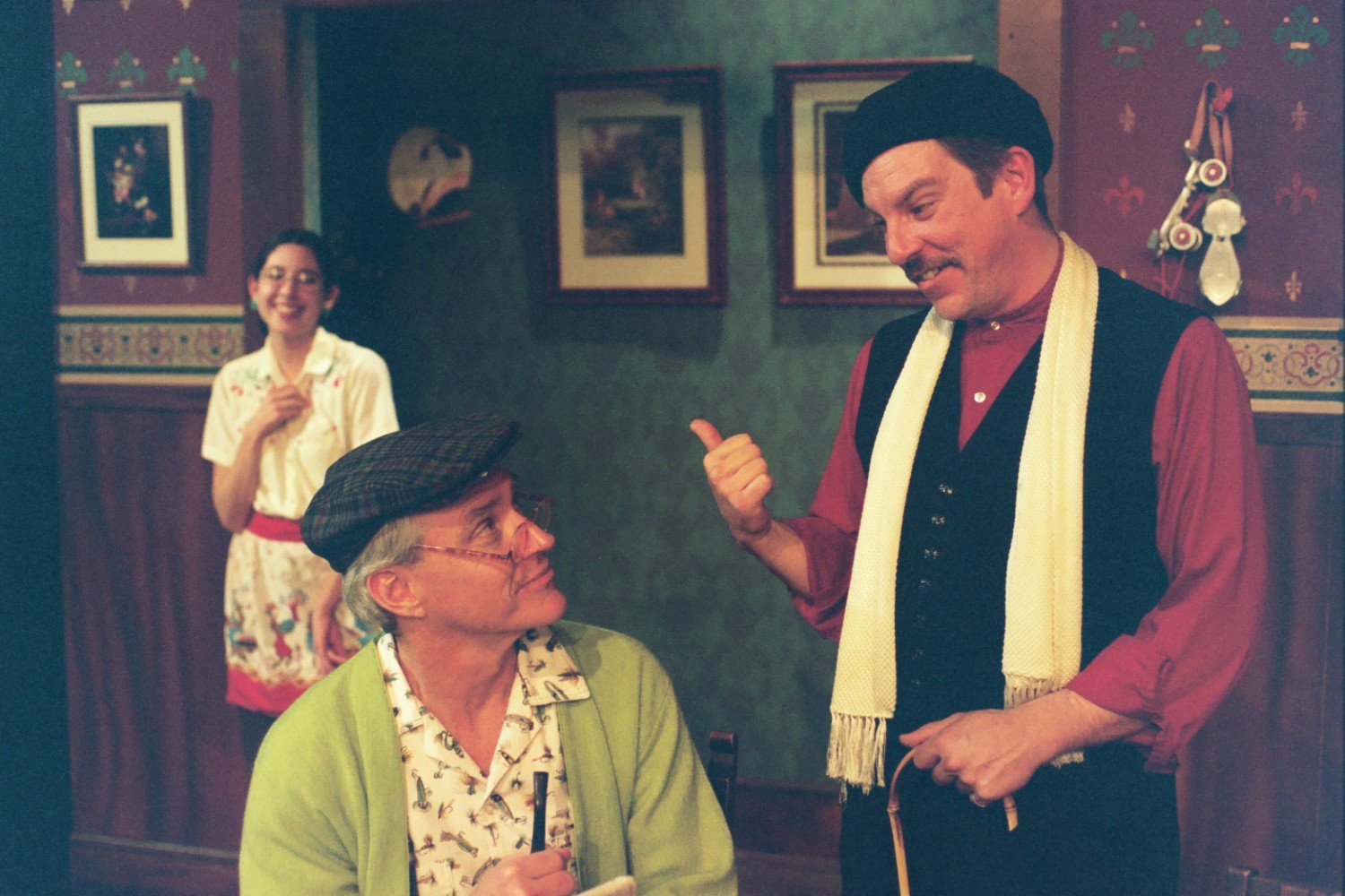 Three people engaged in a theatrical performance; two men are interacting closely, one in a red shirt and black vest with a beret, gesturing with a cane, and the other seated wearing a light green cardigan and cap, holding a pencil and paper. A woman