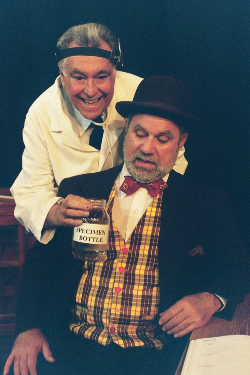 Two men in a theatrical scene; one dressed as a doctor with a head mirror and lab coat, holding a "specimen bottle," and the other wearing a plaid vest, bow tie, and bowler hat, sitting and looking at the bottle.