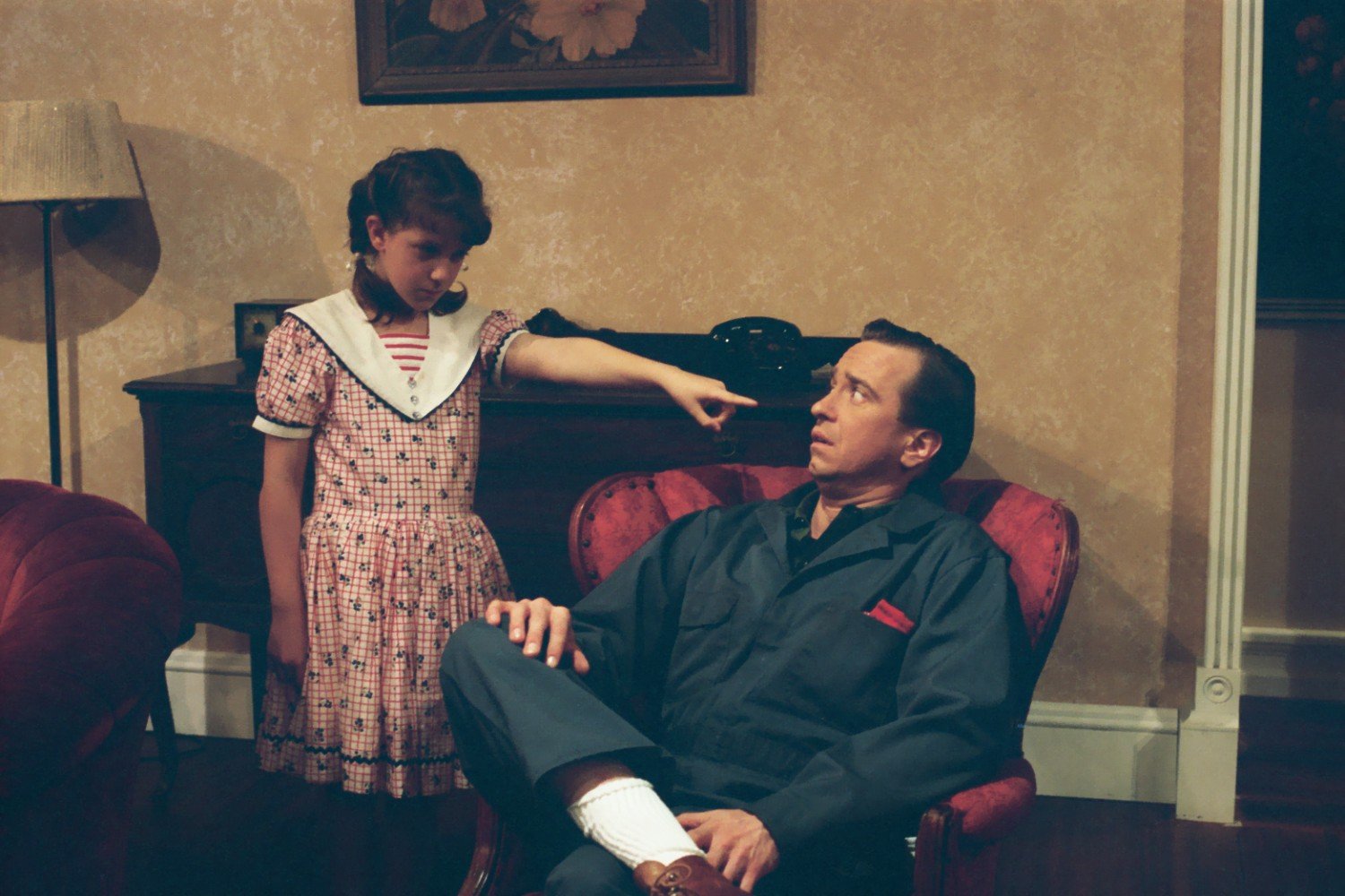 A young girl in a polka dot dress pointing at a seated man who looks surprised. The scene takes place in a vintage-style room with an armchair, a table, and a telephone.