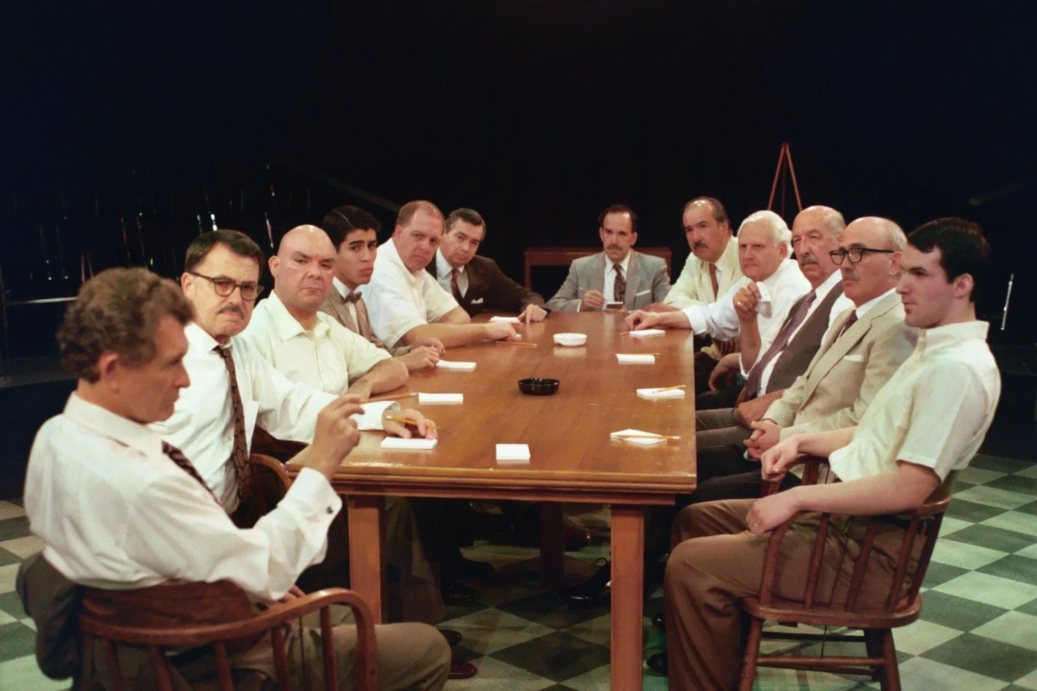 Twelve men in formal attire sitting around a wooden table in a dimly lit room, appearing engaged in a serious discussion.