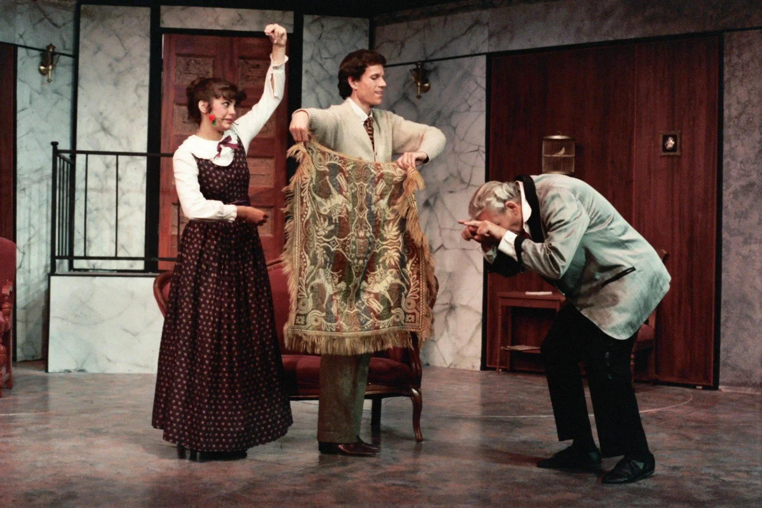 Three actors performing a comedic scene in a theater play. One man is holding a decorative rug, another man is making a humorous gesture, and a woman is playfully posing. The setting includes a marble wall and wooden doors, creating a formal atmosphe