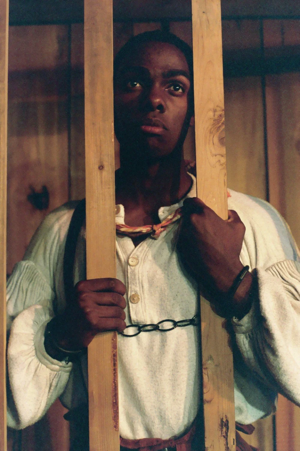 Person in historical attire standing behind wooden bars, wearing handcuffs.