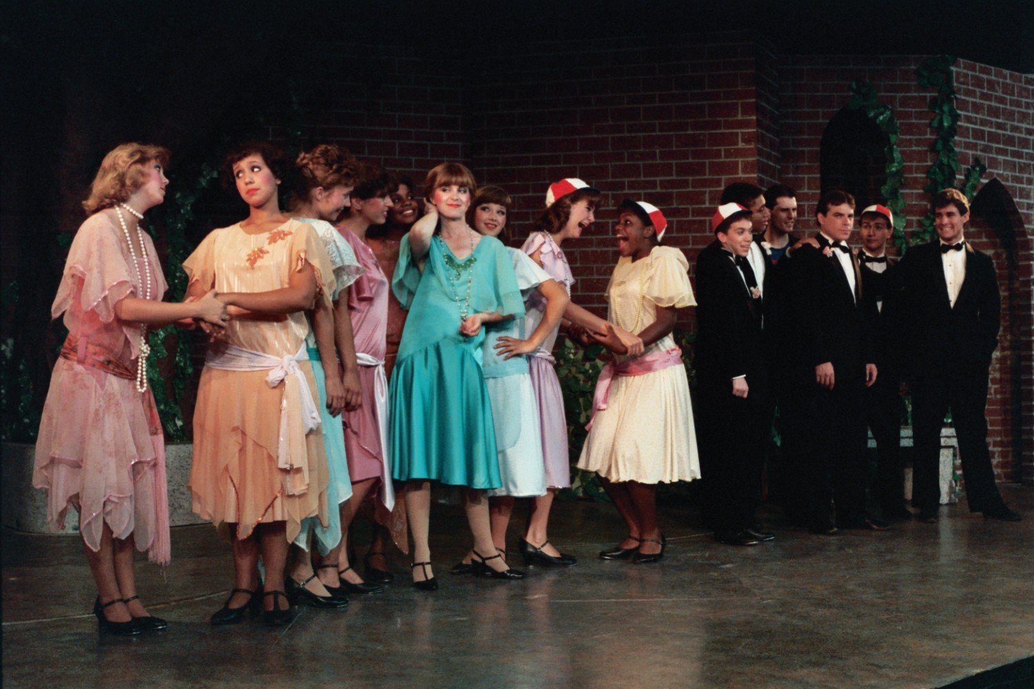 A group of people dressed in formal clothing, with women in pastel dresses and men in tuxedos, stand in a theater setting with a brick wall backdrop.