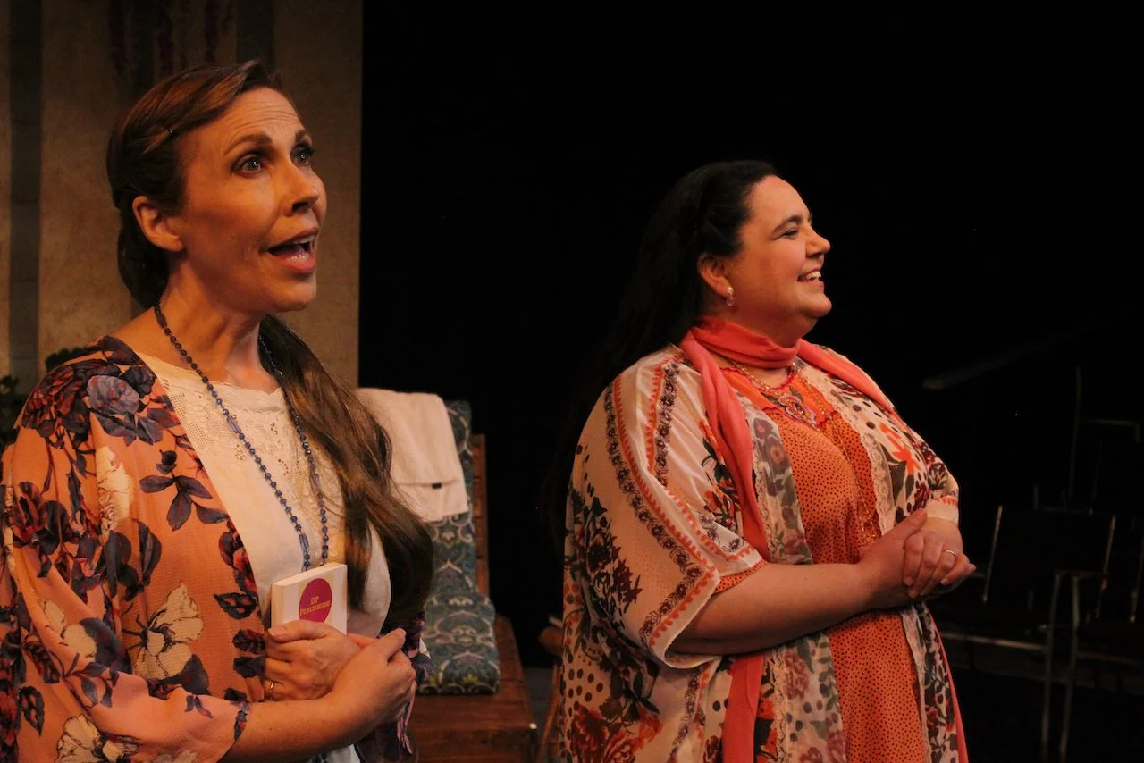 Two women in floral outfits appear on stage, engaged in a conversation, one holding a book.