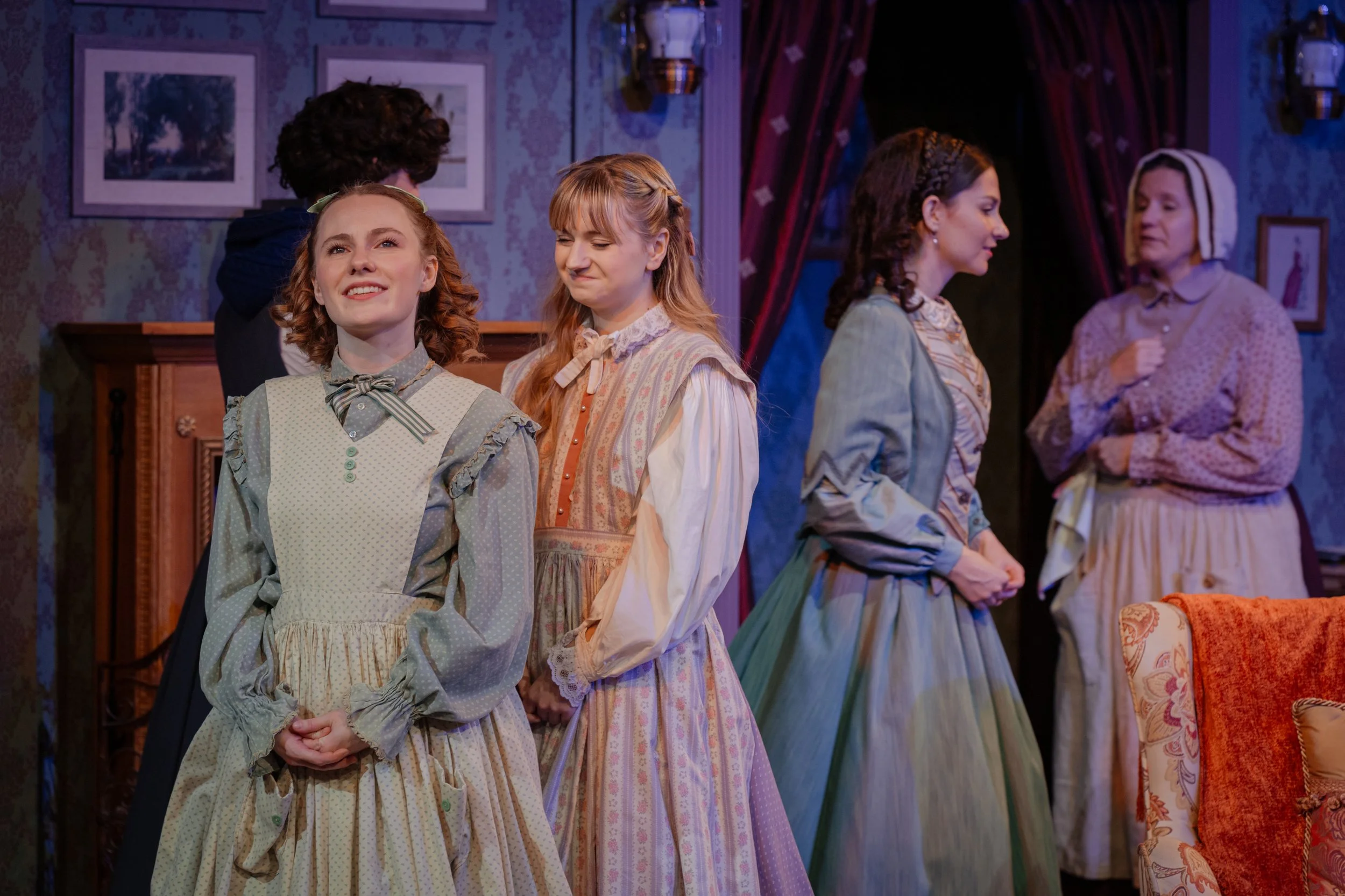 A theater scene with actors dressed in vintage 19th-century attire, including long dresses and aprons, standing on stage in a room decorated with framed pictures and a patterned wall. The setting has theatrical lighting, suggesting a period drama or