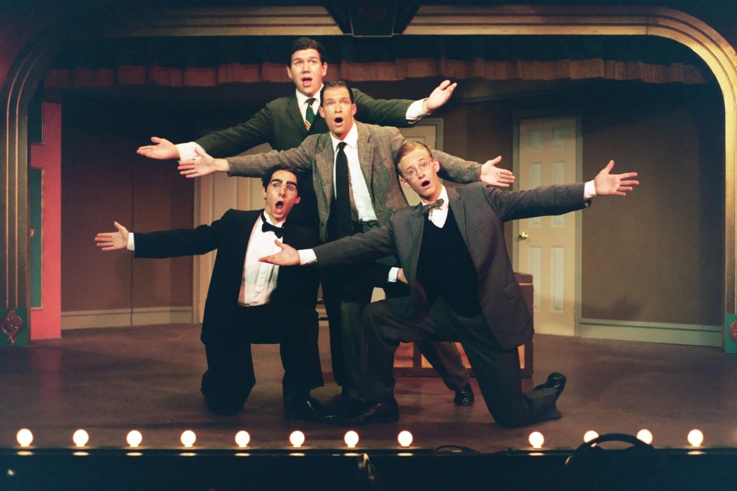 Four men on stage posing dramatically with open arms, dressed in suits and ties, in a theater setting with stage lights and curtains in the background.