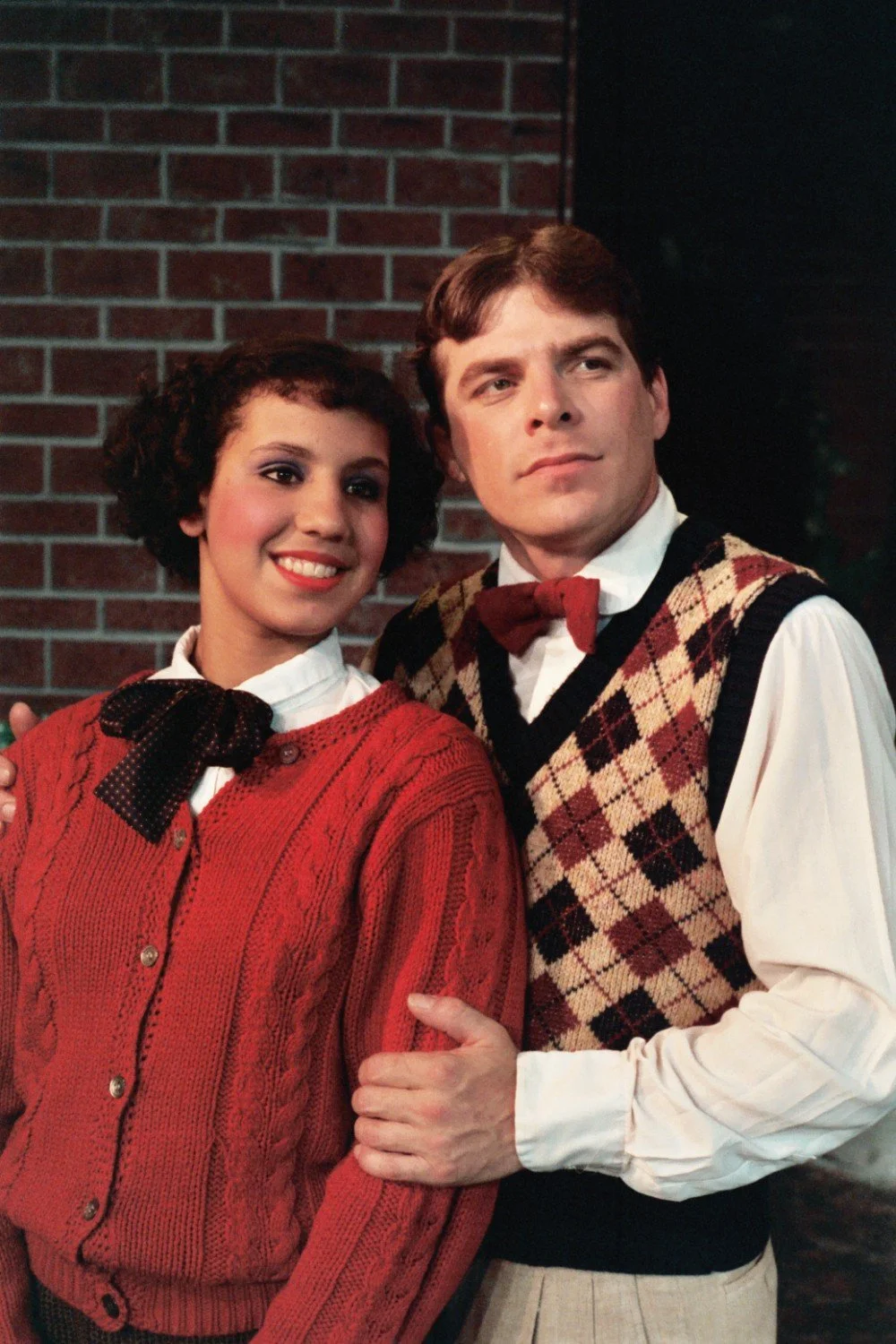 A man and woman dressed in vintage attire standing in front of a brick wall. The woman is wearing a red sweater with a polka dot bow, and the man is wearing a bow tie and argyle sweater vest.