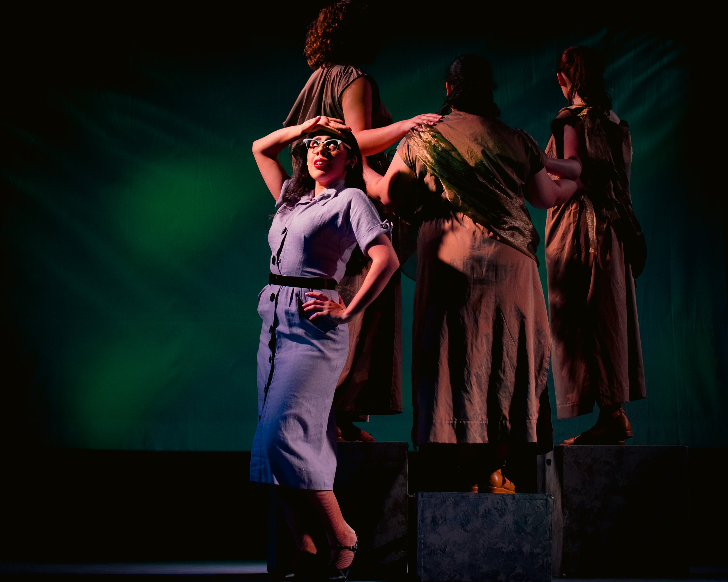 A theatrical performance with a person in the foreground wearing sunglasses and a light blue dress. They are striking a dramatic pose with one hand on their hip and the other on their head. Four figures in the background are wearing earth-toned costu