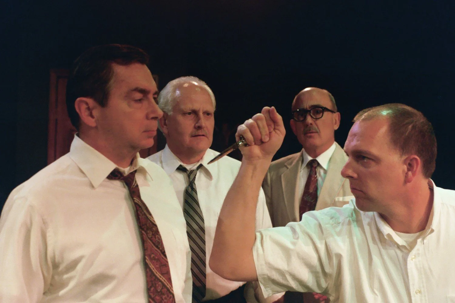 Four men in formal attire, with one holding a pointed object, possibly in a tense discussion or dramatic scene, against a dark background.