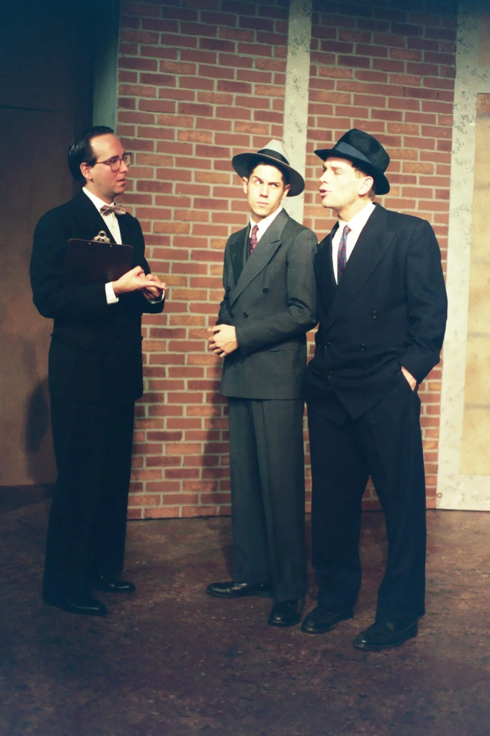 Three men in vintage suits and hats having a conversation in front of a brick wall, possibly part of a theater production.