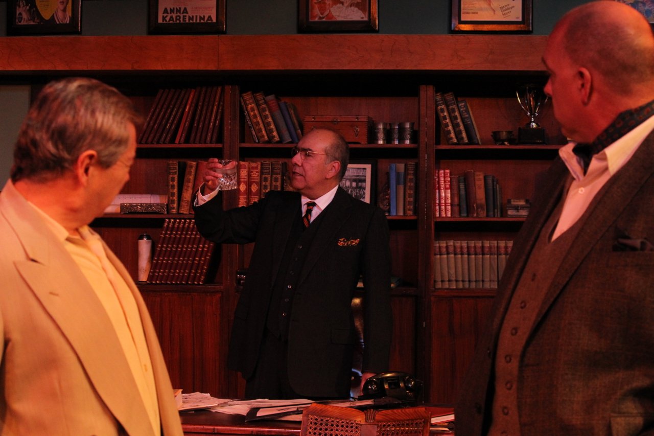 Three men in a room with bookshelves, one holding a glass, having a conversation, dressed in suits, wooden desk with papers.