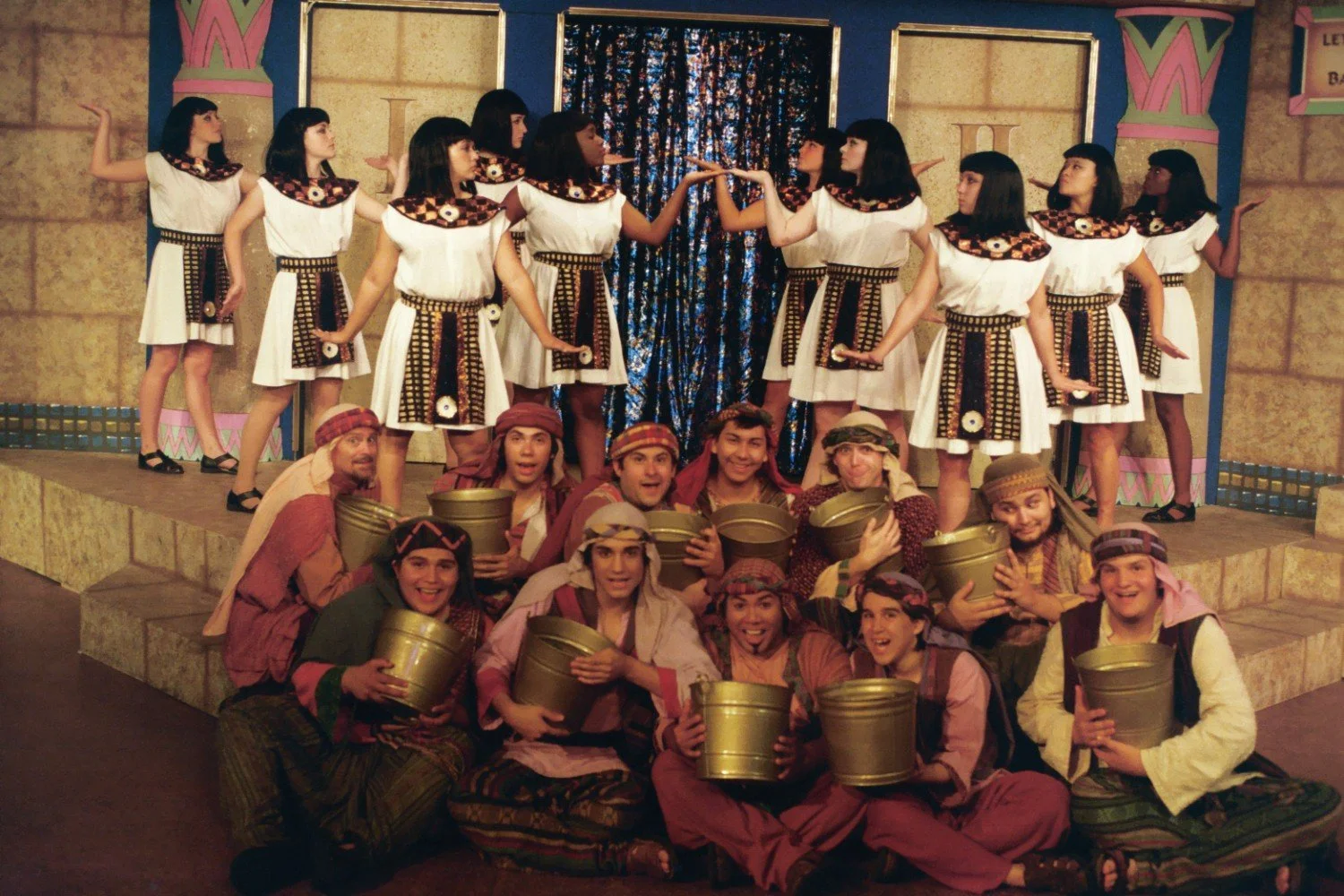 Group of performers in theatrical costumes, some dressed as ancient Egyptians with white dresses and geometric collars, others as Middle Eastern characters with headscarves and robes, holding buckets, on a stage with a stone backdrop and columns.