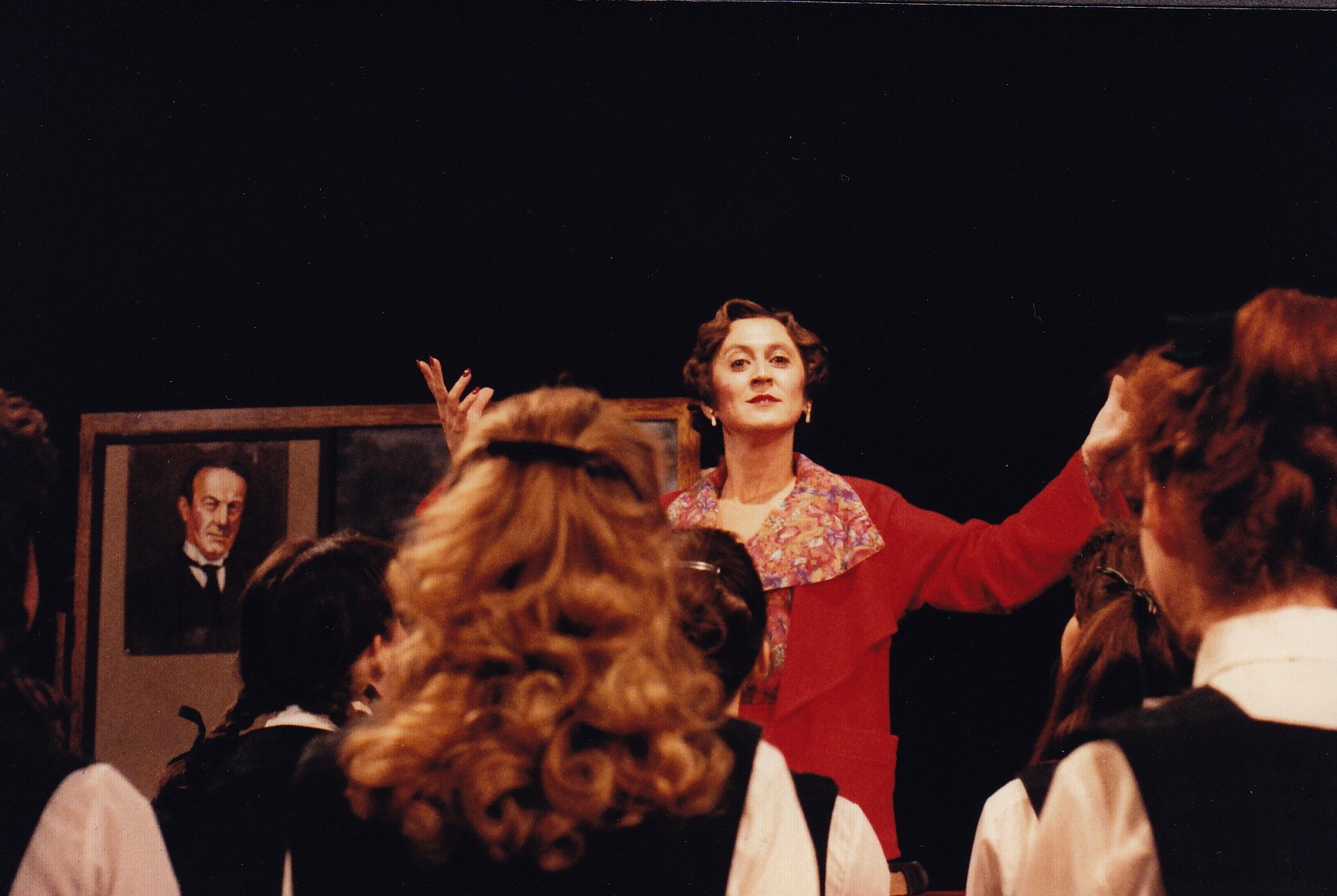 A woman in a red outfit standing in front of a group of people with a portrait in the background.