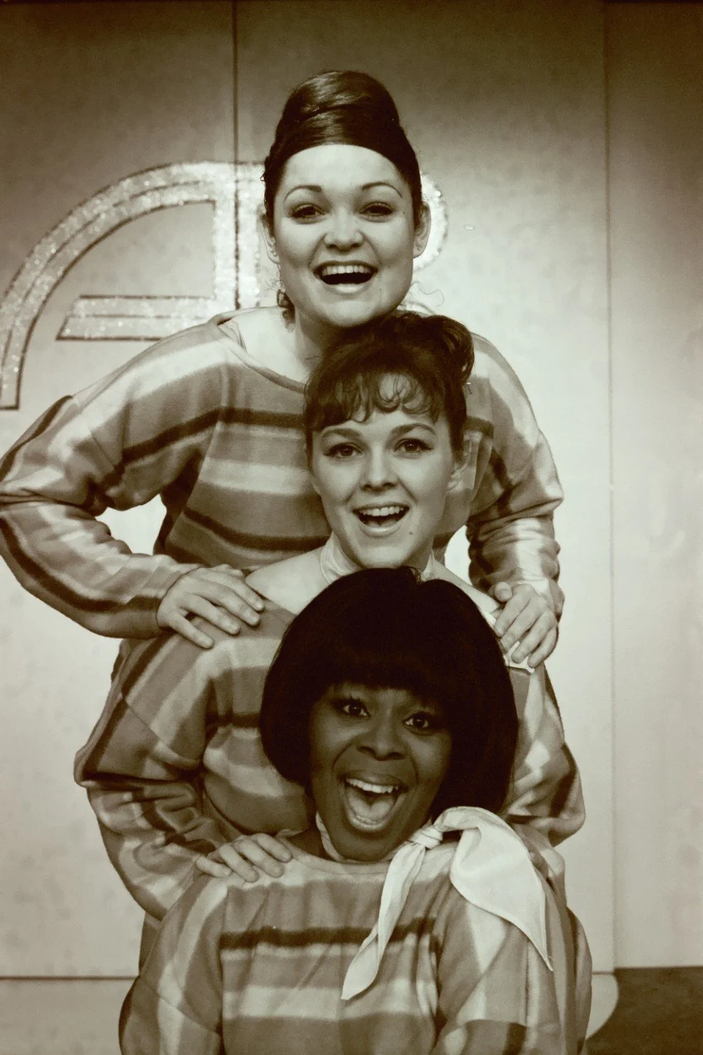 Three women smiling, dressed in matching striped outfits, in a stacked pose.