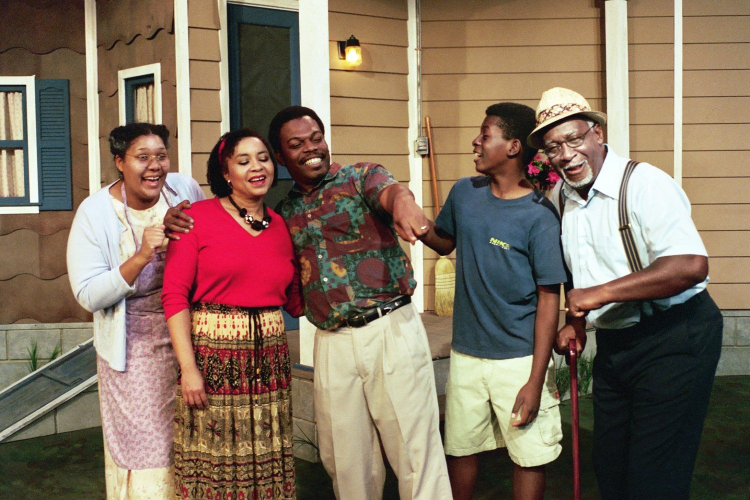 Group of diverse people laughing and pointing in front of a house with a porch.