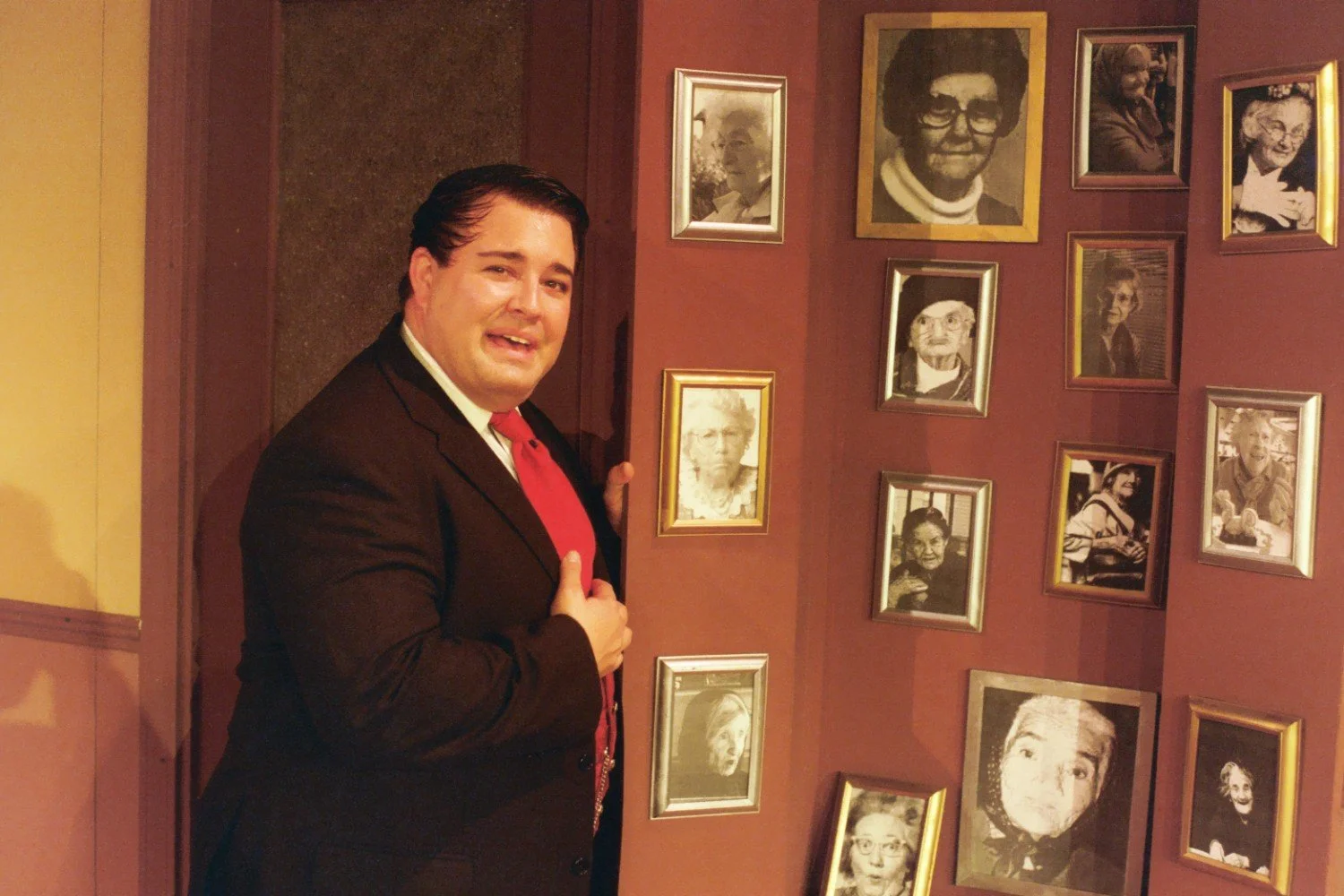 A man wearing a suit and red tie stands in front of a wall with multiple framed photographs. The photographs are black and white portraits of various elderly people.