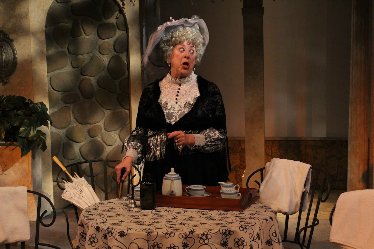 A person in period costume with a bonnet, standing at a table with tea set and umbrella in a decorated room.