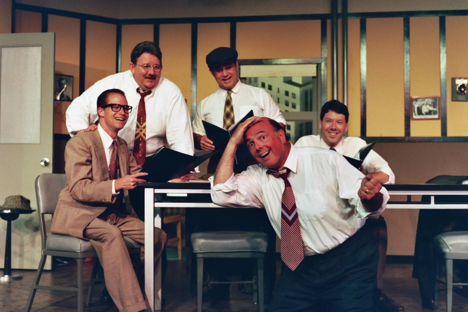 Group of five men in an office setting, laughing and holding papers, with a comedic pose from one man.
