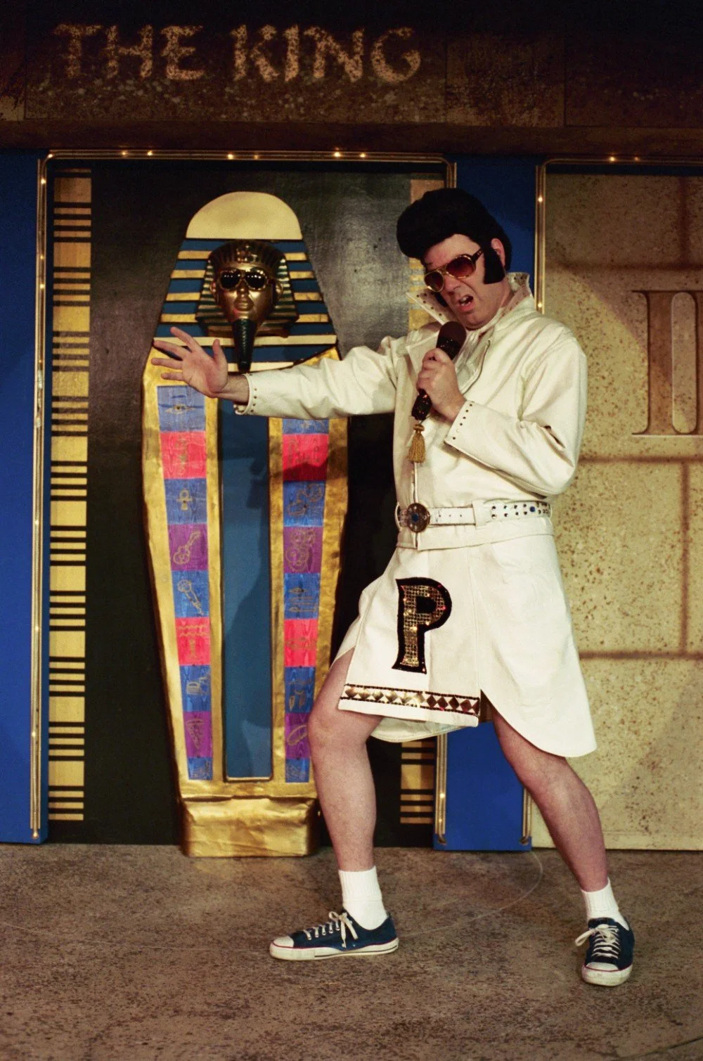Person dressed as Elvis Presley impersonator, holding microphone, standing in front of Egyptian-themed backdrop.