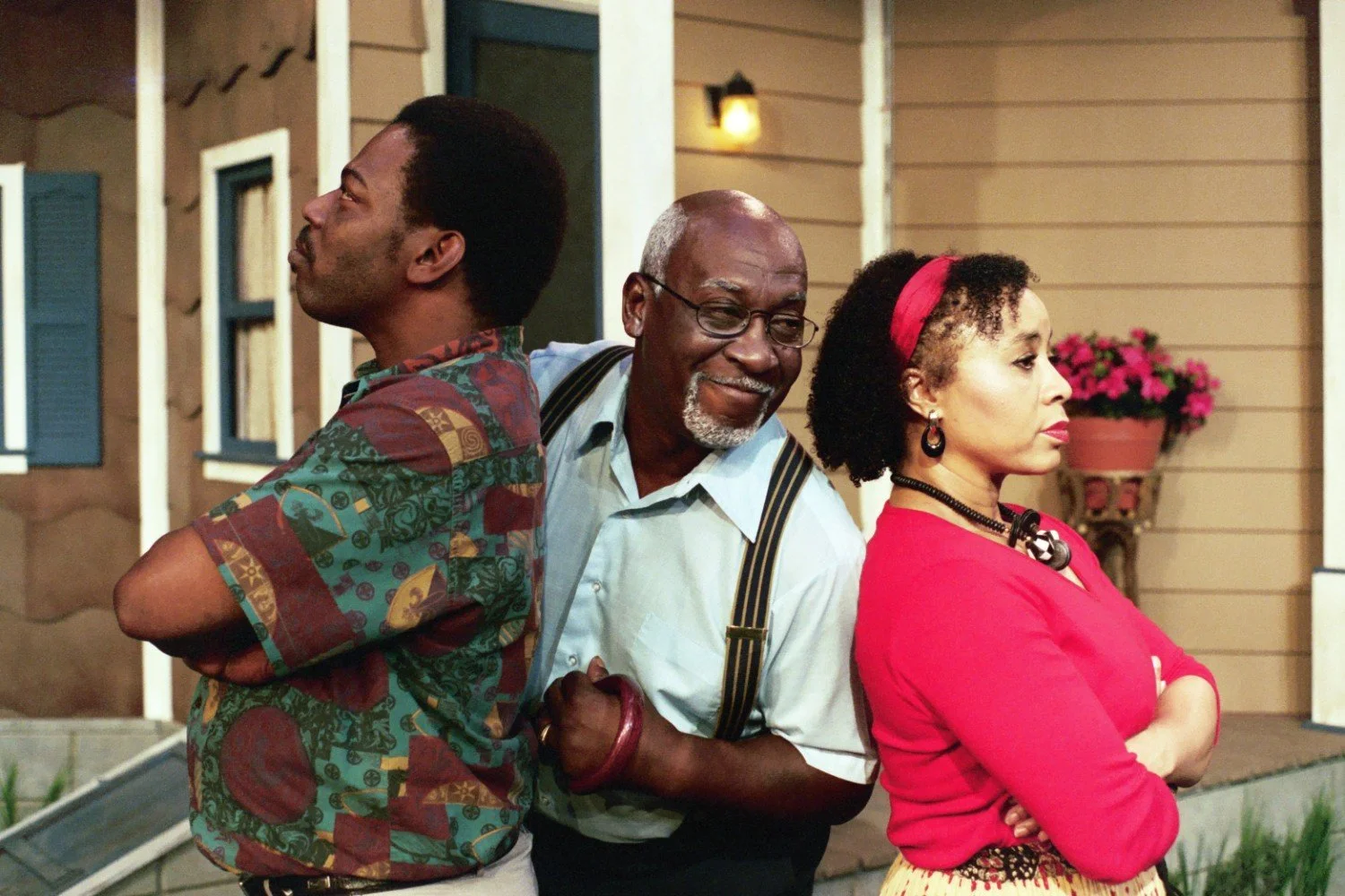 Three people standing in front of a house, with two of them back to back looking away, and the third smiling in the middle.