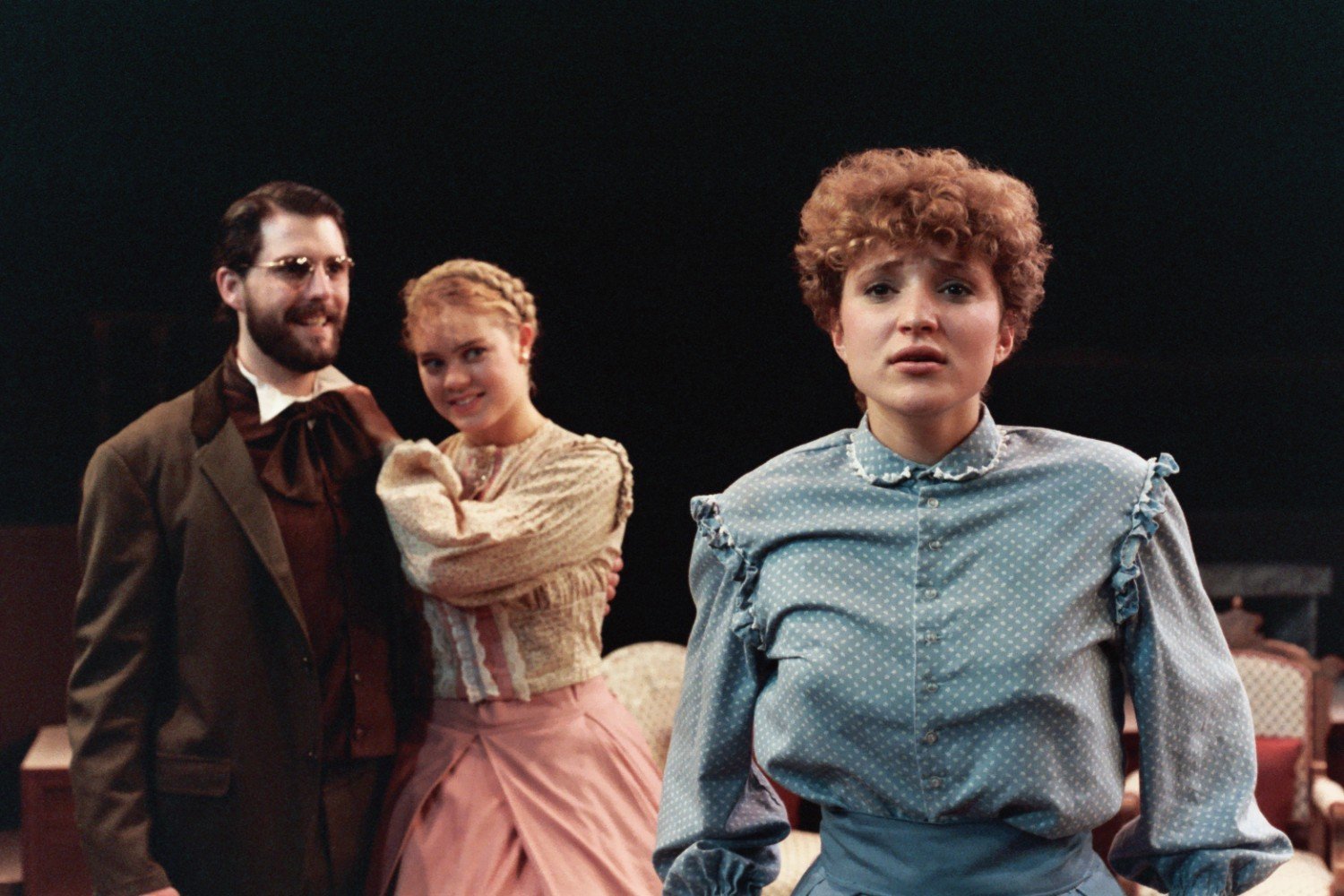 Three actors in period costumes performing on stage, with one woman in the foreground looking emotional, and a man and woman in the background smiling.