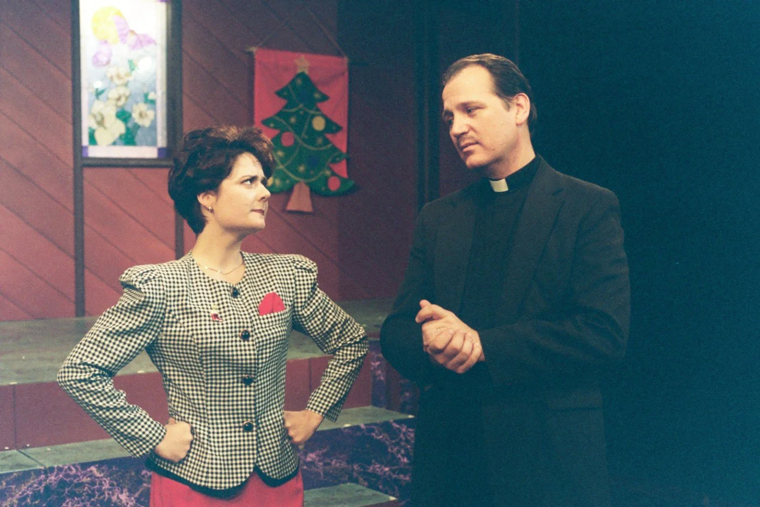 A woman in a checkered suit and a man dressed as a priest are having a conversation on a stage set with a Christmas tree banner and floral artwork.