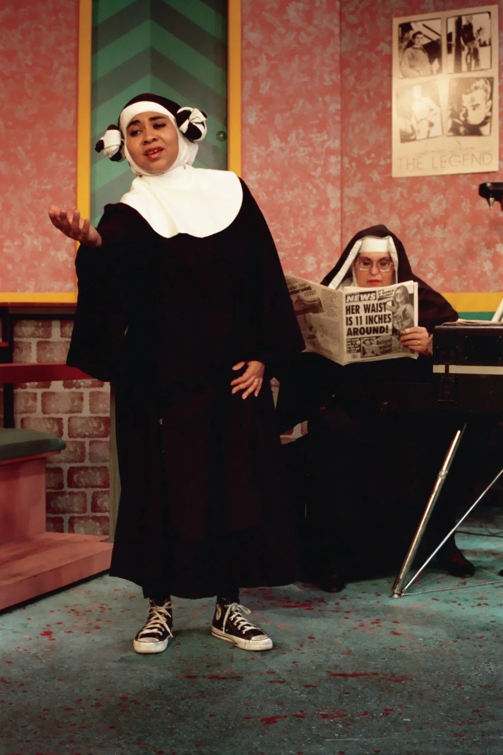 Two people dressed as nuns in a comedic setting; one is singing with rolled hair and sneakers, the other reads a newspaper with headlines.