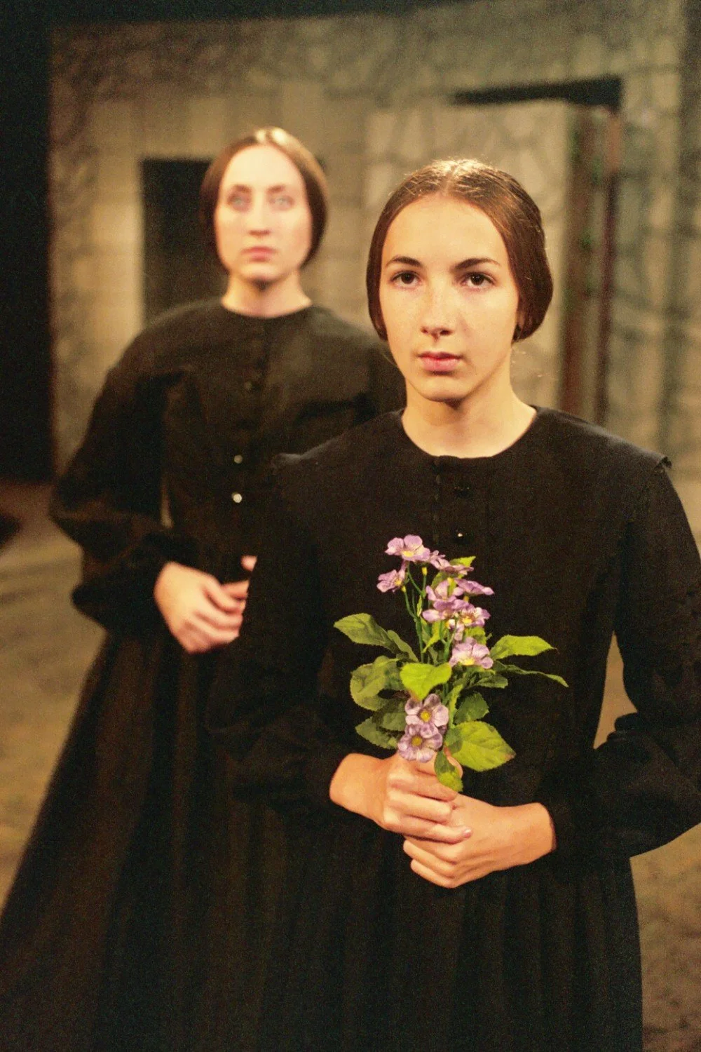 Two women in black dresses, with one holding purple flowers, set against a stone background.
