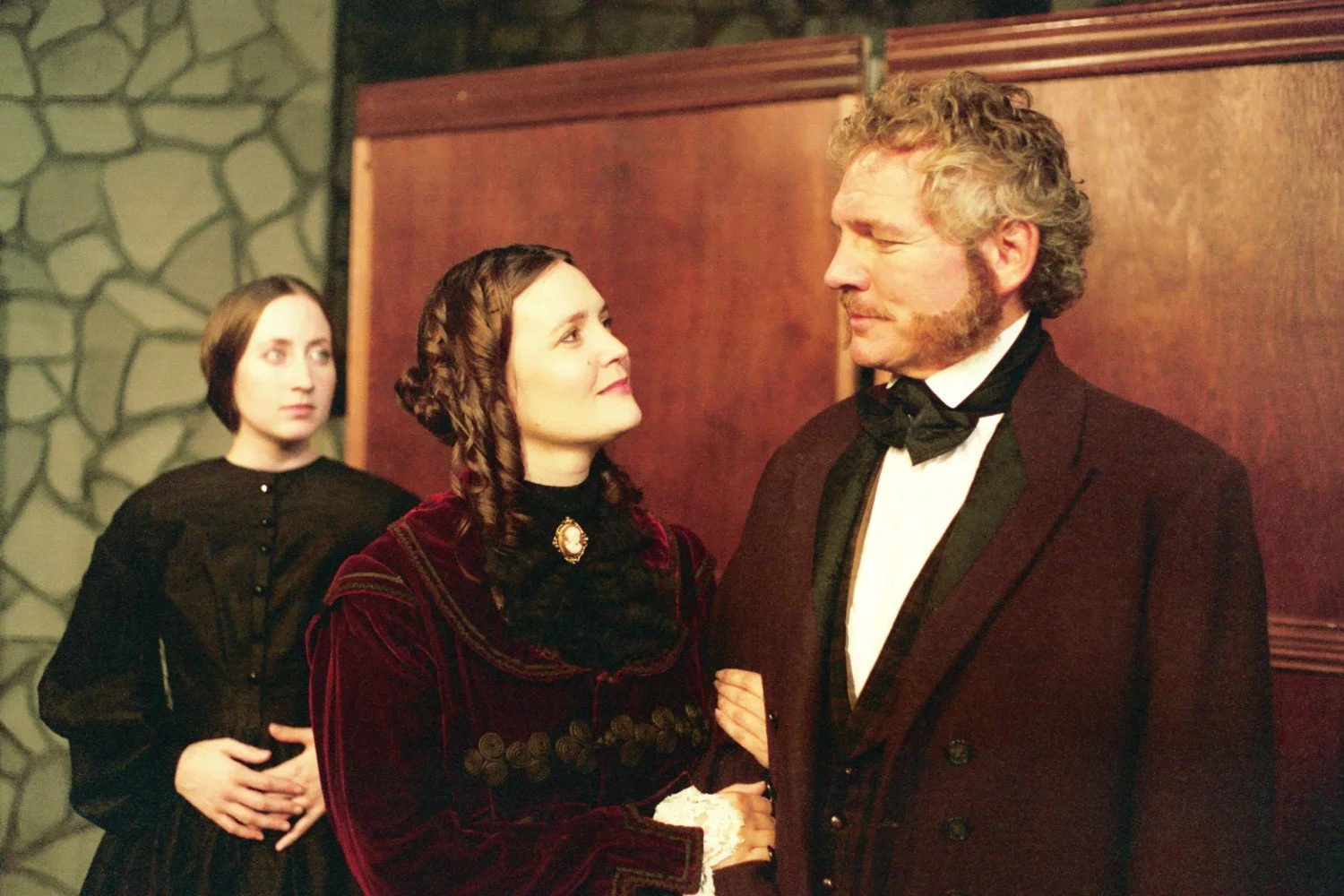 Three people in vintage 19th-century attire. A woman in a dark dress and lace collar holds the arm of a man with curly hair and sideburns, wearing a suit and bow tie. Another woman in a simple dark dress stands slightly behind them, with a stone wall