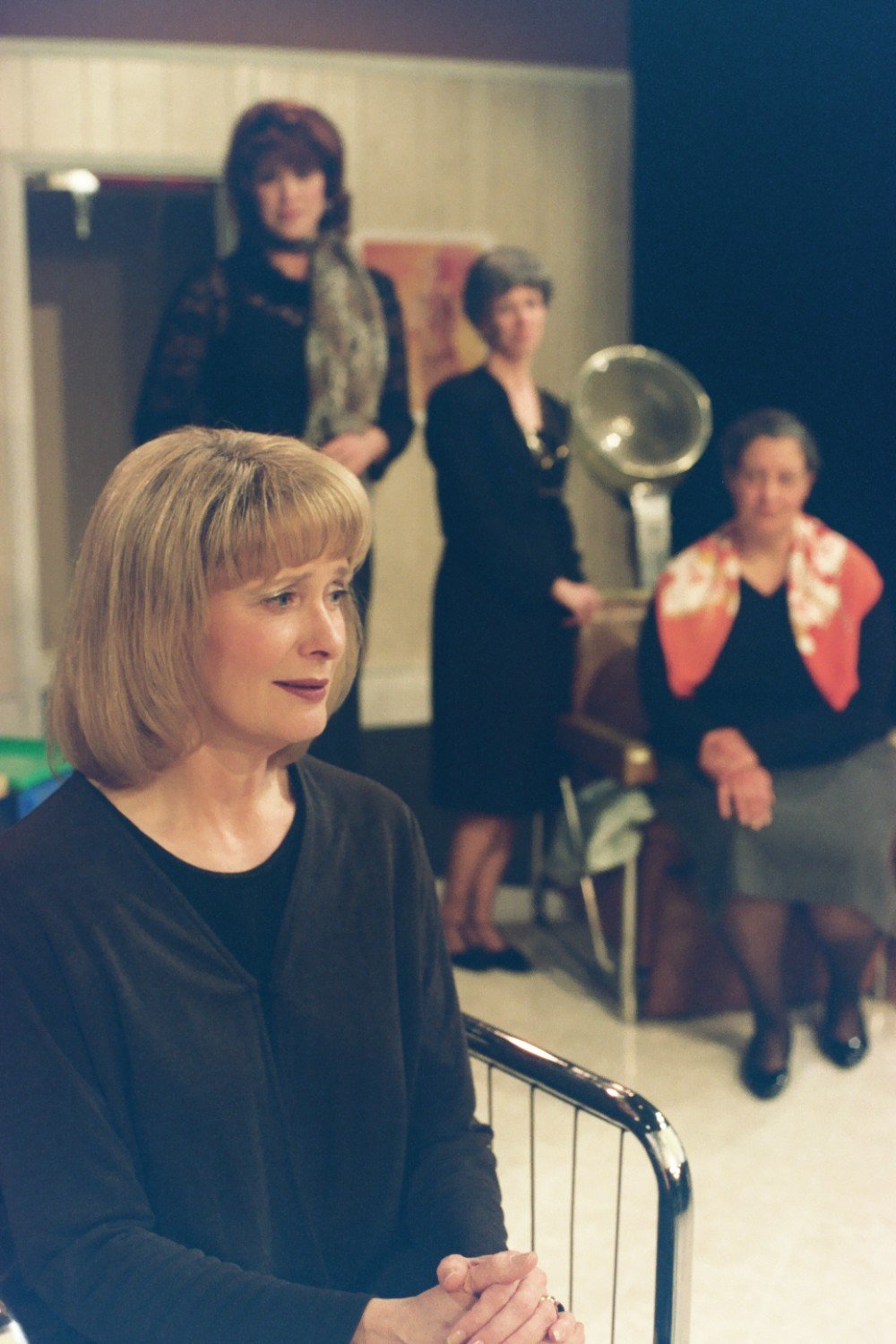 Four women in a room, one sitting in front, the others standing and sitting in the background, with a vintage hair salon setting.