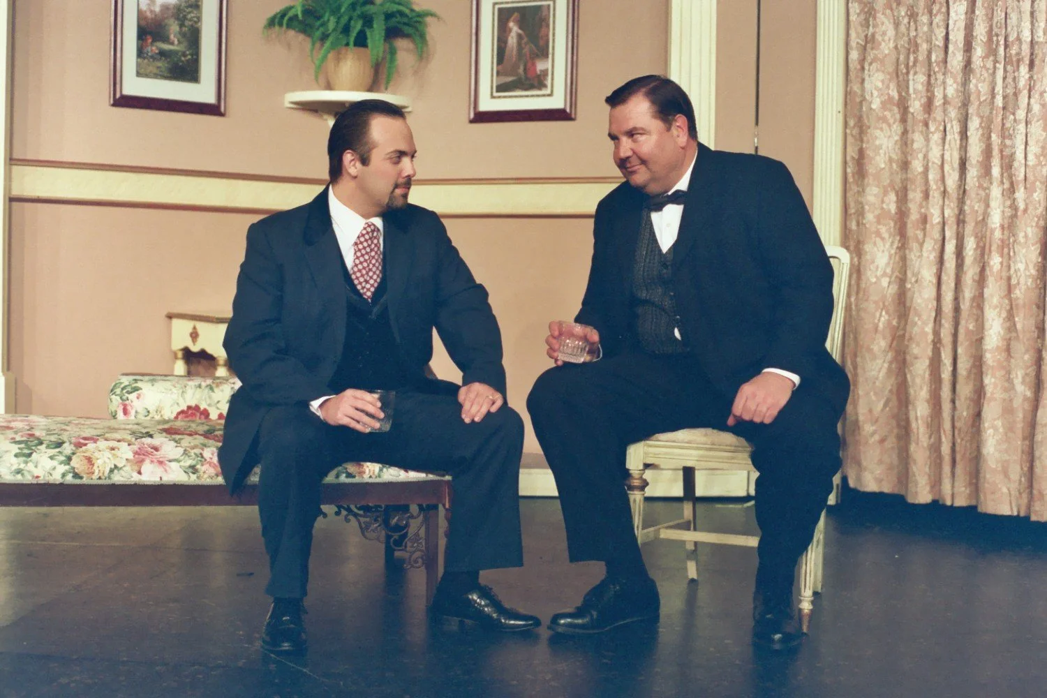 Two men in suits conversing in a vintage-style room, holding drinks; one is seated on a floral bench, the other on a chair.