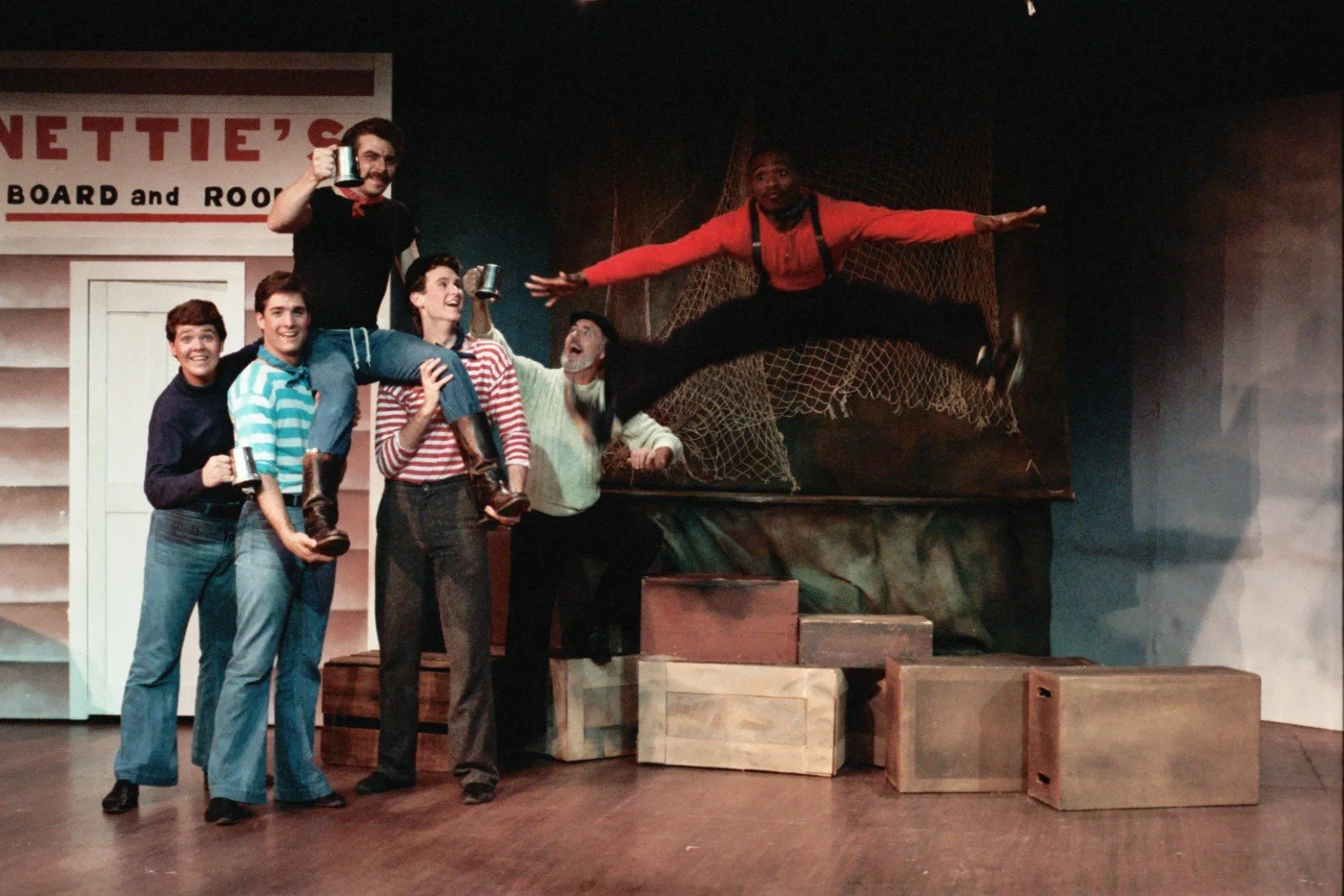 Group of actors performing on stage, one performing a split jump, others holding metal mugs and lifting a person; set includes crates and a fishnet backdrop.