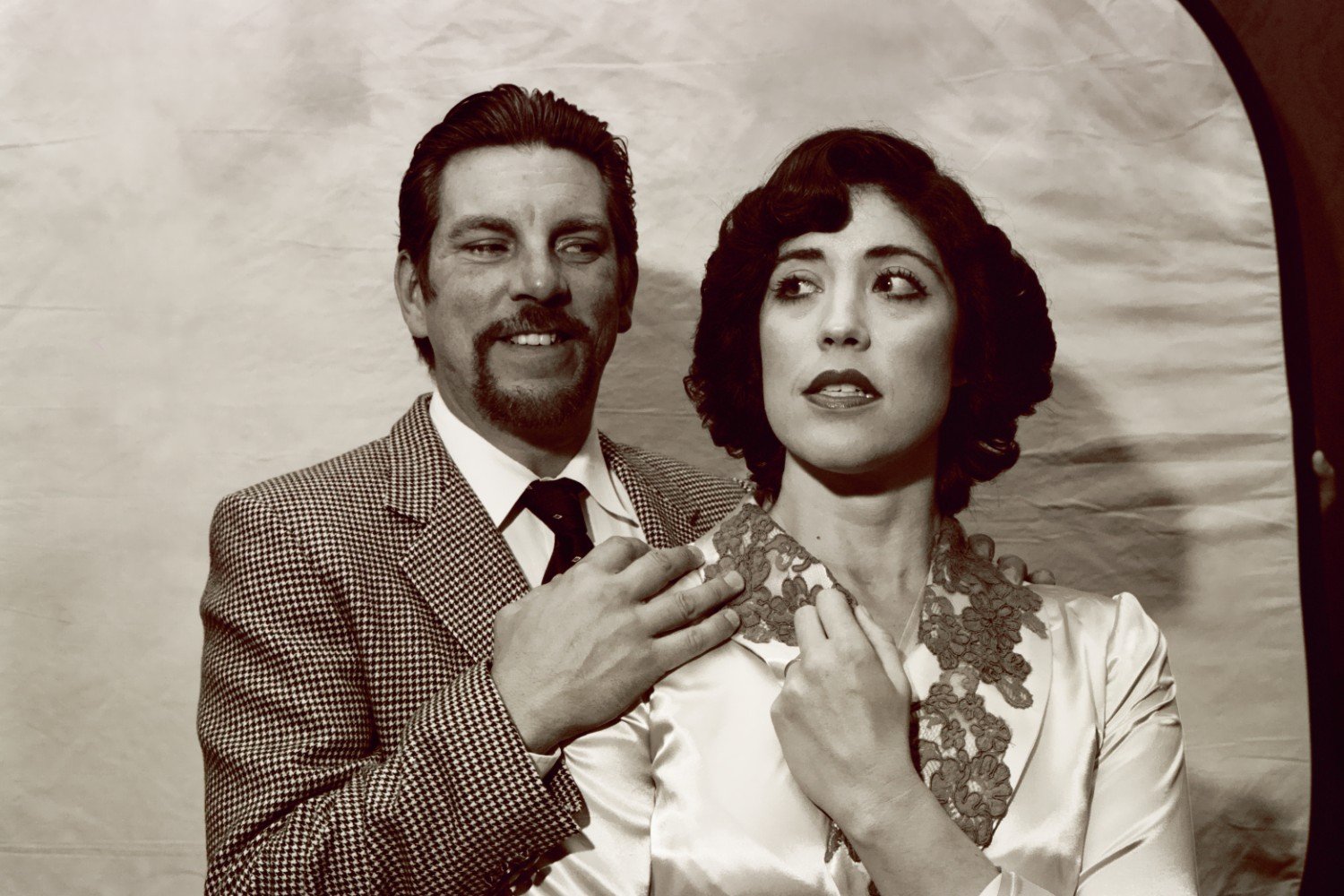 Sepia-toned image of a man in a suit smiling and standing behind a woman in a vintage dress, both in a posed, dramatic setting.