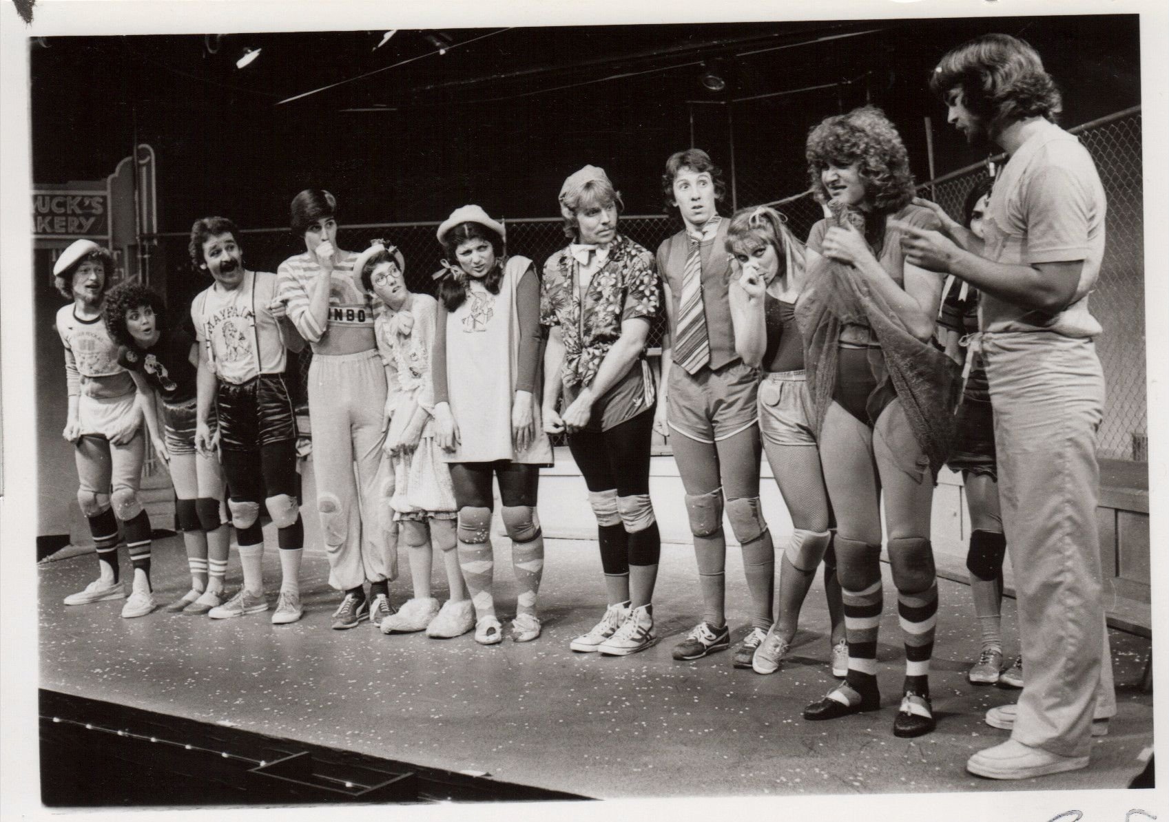A group of people dressed in casual and athletic attire stand in a line on a stage, appearing to be part of a theatrical performance. They are wearing a mix of shorts, knee pads, and shirts. The stage setting includes a backdrop with a sign advertisi