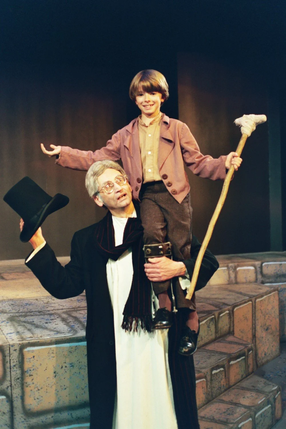 A man in period costume holding a boy with a crutch on stage