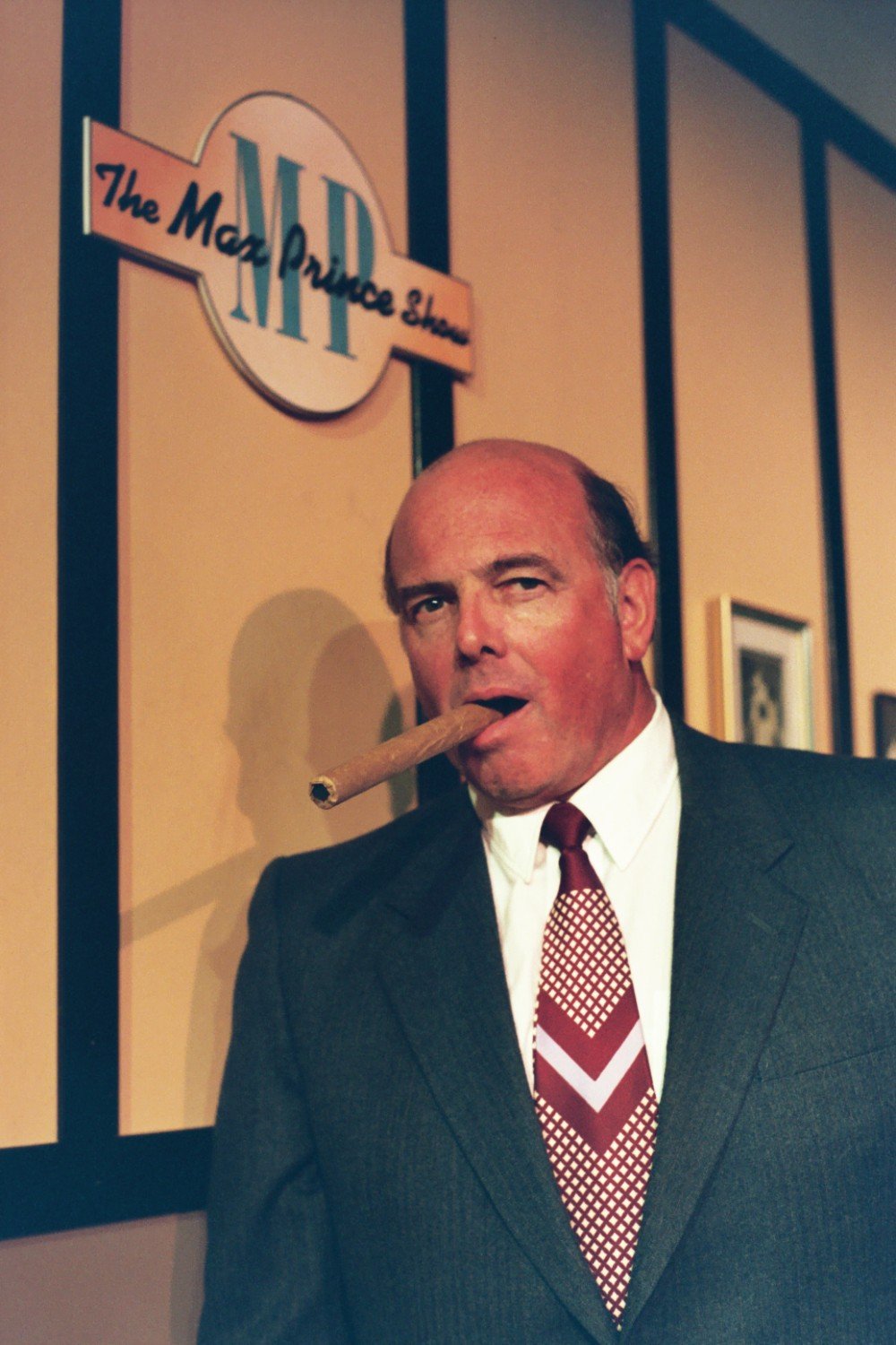 Man in a suit with a cigar, standing in front of "The Max Prince Show" sign.