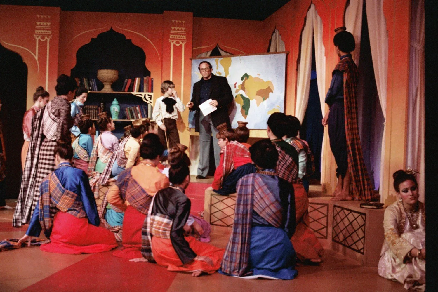 Scene from a theatrical performance with actors in colorful costumes, gathered around a man in front of a map and a bookshelf, set in an ornate room.