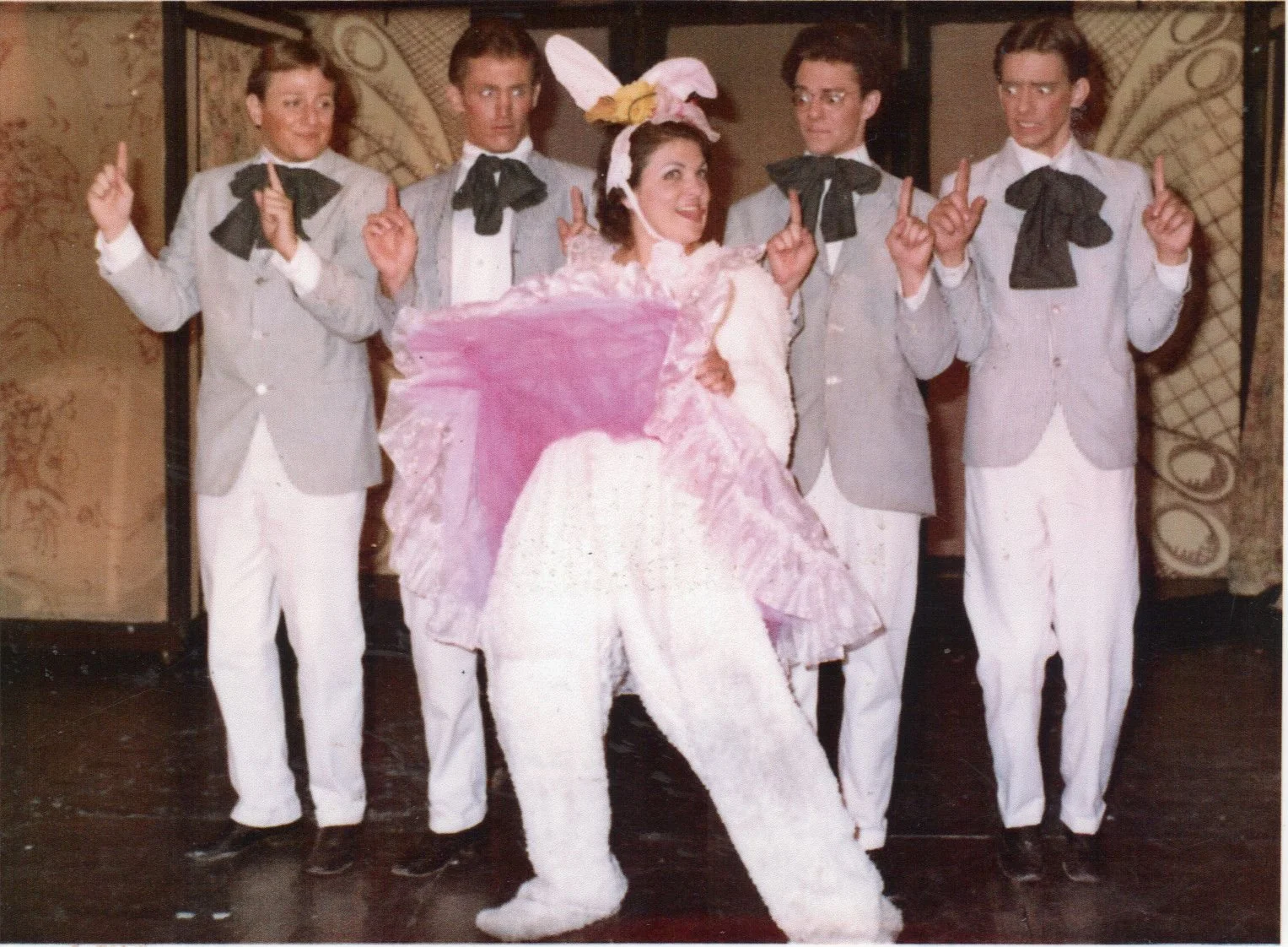 A group of four men in gray suits with black bow ties posing with a woman in a pink and white bunny costume on a stage, performing a theatrical act.