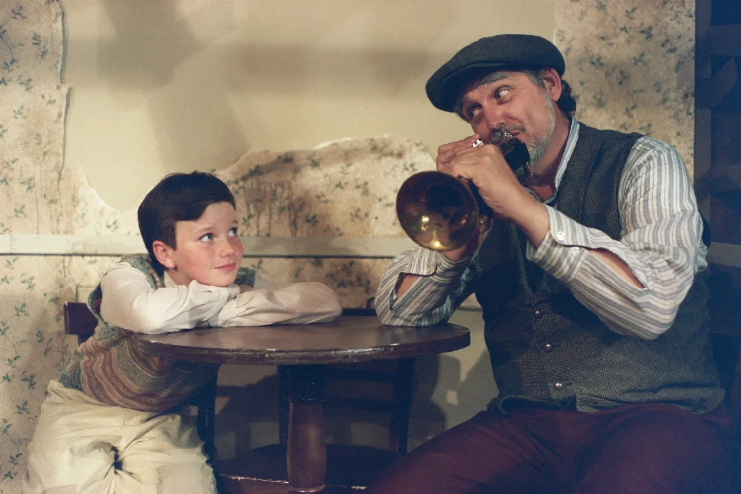 A young boy sitting at a table, leaning forward with arms crossed, gazing at an older man playing a horn, who is wearing a flat cap and vest.