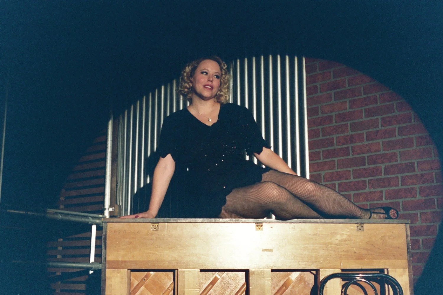 Woman in black dress sitting on piano in dimly lit room with brick and metal background.