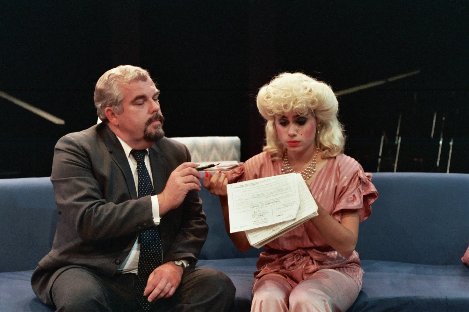 A man in a suit hands a pen to a woman with blonde curly hair in a pink outfit, seated on a blue couch, holding paperwork.