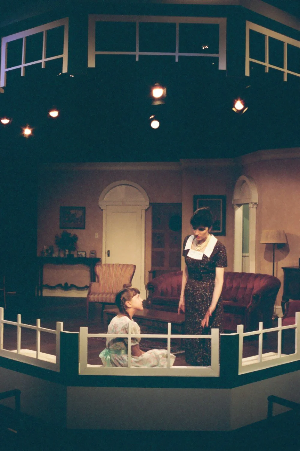 Two actors on a theater stage set resembling a living room, with a woman standing and a girl sitting on a low platform. The room is decorated with vintage furniture and props, including a sofa, chair, and table.