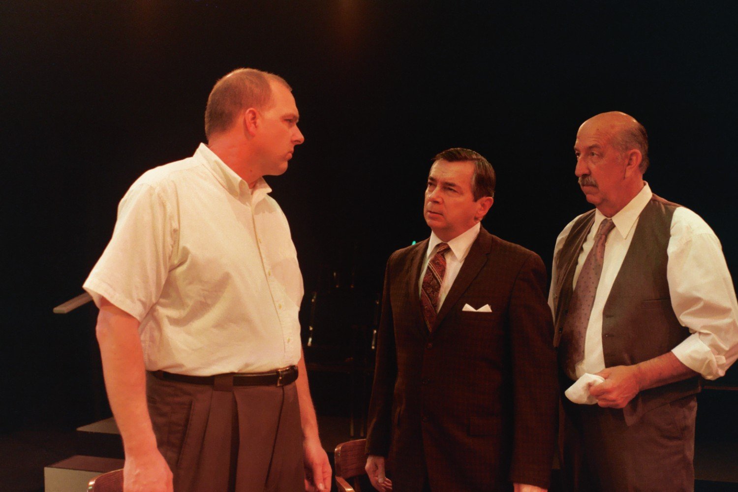 Three men engaged in a serious conversation on stage, dressed in formal attire, with one wearing a shirt, another in a suit, and the third in a vest.