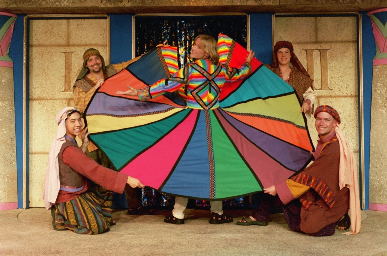 Performers in colorful costumes on stage, highlighting a vibrant, multicolored coat as part of a theatrical performance.