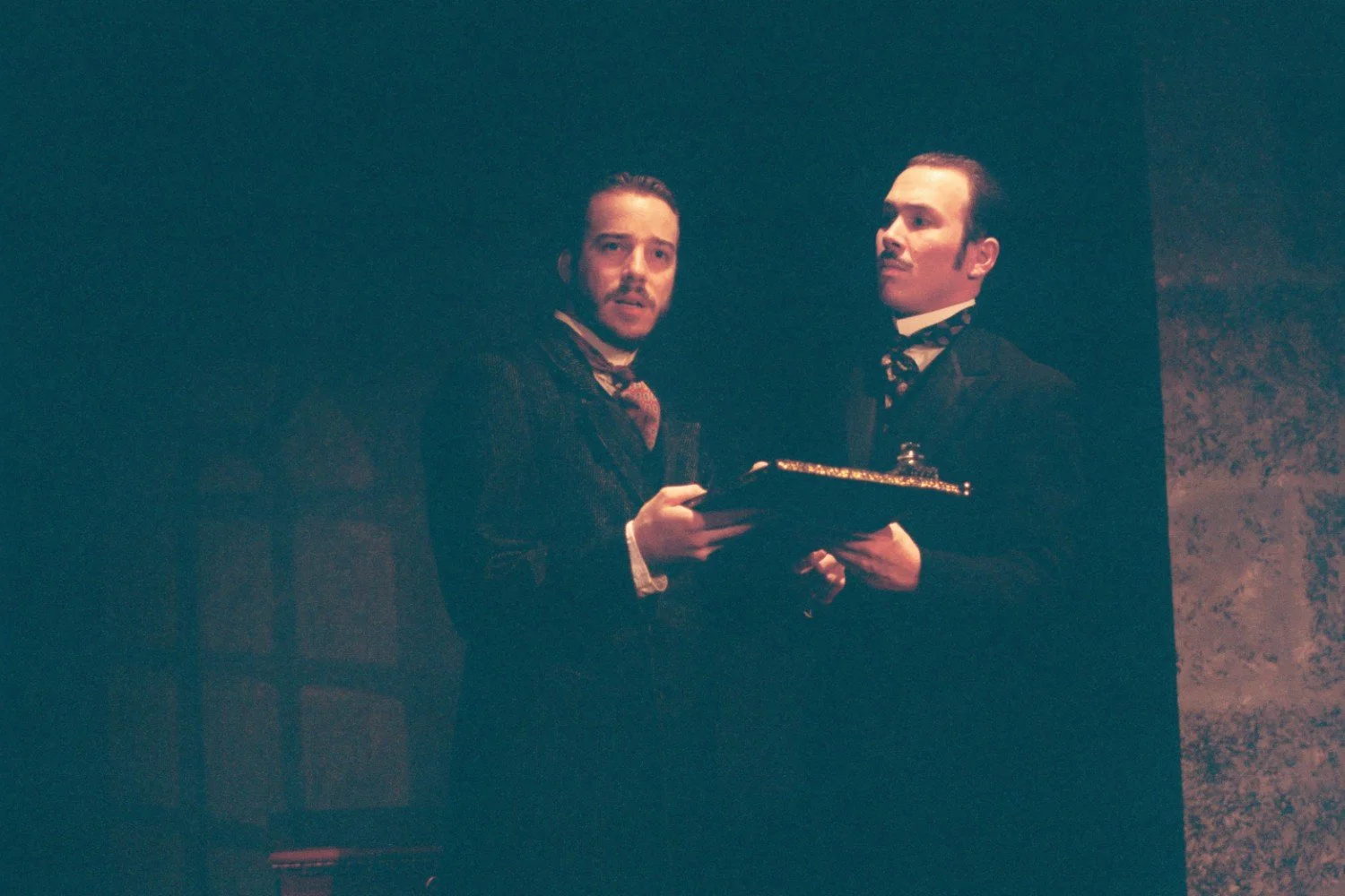 Two men in period costumes onstage, holding a framed item, under dramatic lighting.