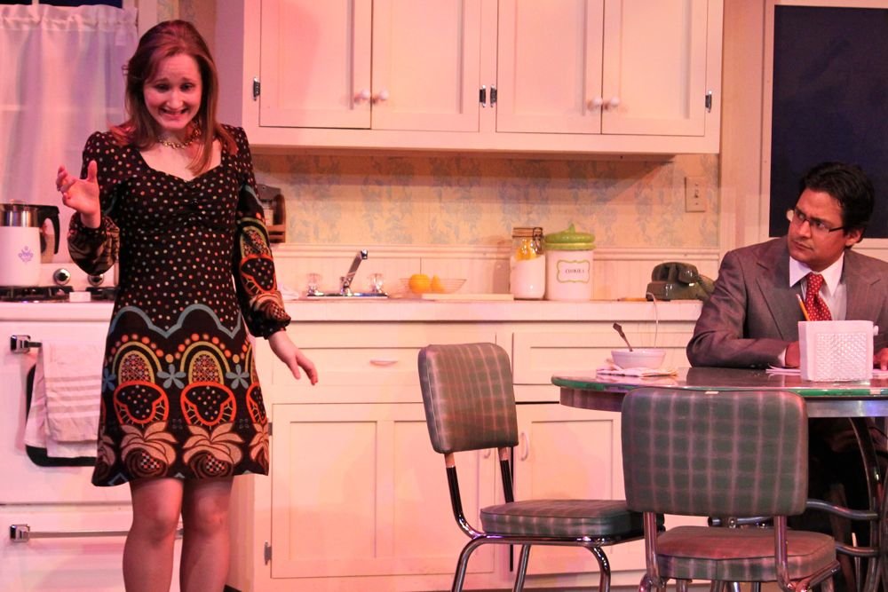 A woman in a colorful patterned dress stands in a kitchen with a concerned expression. She is gesturing with one hand. A man in a suit and tie sits at a table with a cup and papers, looking at her intently. The kitchen features white cabinets, a stov